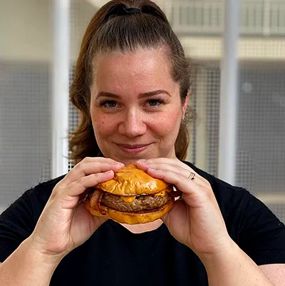 Amanda Magalhães sorrindo e segurando um hambúrguer gourmet pronto para ser saboreado. Ela está em um ambiente casual com tijolos à vista, transmitindo uma atmosfera acolhedora e descontraída, assim como a do Curso Completo do Hambúrguer Perfeito!