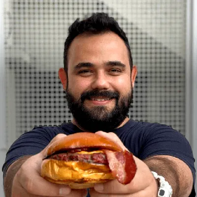 Fabricio Schibuola em close-up, oferecendo um hambúrguer artesanal com um sorriso convidativo. O fundo desfocado sugere um ambiente ao ar livre, destacando o hambúrguer como o foco da imagem. Venha participar do Curso Completo do Hambúrguer Perfeito!